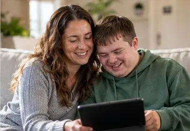 A mother and her adult son smiling at a tablet together on the couch