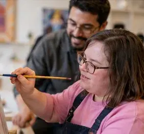 A woman painting at an easel while a support person looks on