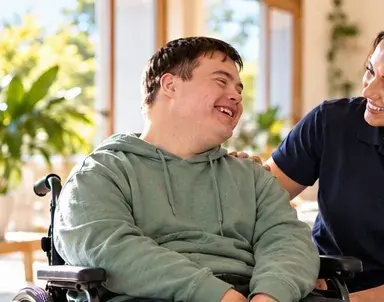 A young man in a wheelchair laughing with a support worker in a bright living room